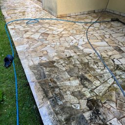 Pozamiatane - Czyszczenie kostki brukowej na tarasie z użyciem myjki ciśnieniowej. Widoczna różnica w czystości między umytą a brudną częścią powierzchni. Wąż ogrodowy na trawie.
