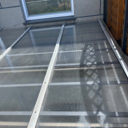 Pozamiatane - Przezroczyste zadaszenie z poliwęglanu, wsparte na metalowej konstrukcji, przylegające do ściany budynku z oknem. Widoczny fragment ogrodzenia.