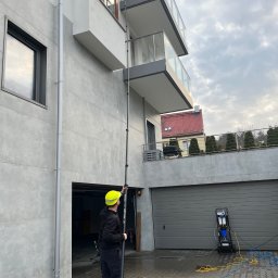 Pozamiatane - Pracownik w kasku myje elewację domu nowoczesną lancą teleskopową. Widoczny sprzęt do mycia ciśnieniowego i przewody. Szara elewacja, balkony z barierkami.