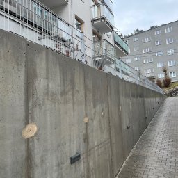 Pozamiatane - Szara, betonowa ściana oporowa z widocznymi śladami po naprawach, przylegająca do chodnika z kostki brukowej, z budynkiem mieszkalnym w tle. Ujęcie z dołu.
