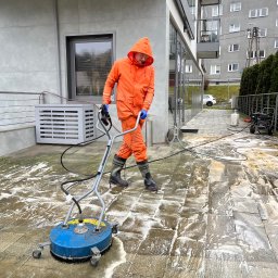 Pozamiatane - Pracownik w pomarańczowym kombinezonie czyści kostkę brukową na zewnątrz budynku za pomocą specjalistycznej maszyny ciśnieniowej. Widoczne zabrudzenia i piana.