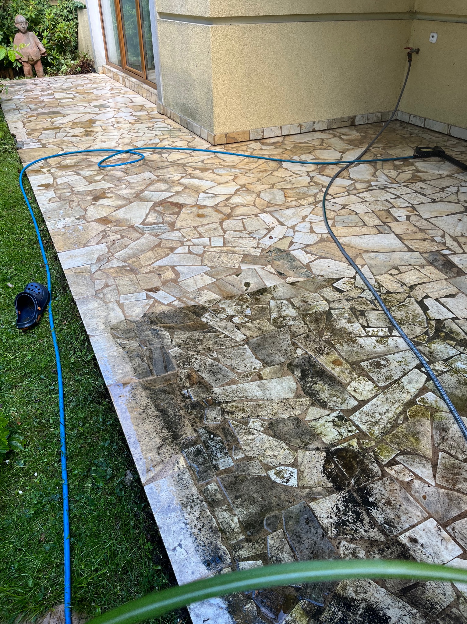 Czyszczenie kostki brukowej na tarasie z użyciem myjki ciśnieniowej. Widoczna różnica w czystości między umytą a brudną częścią powierzchni. Wąż ogrodowy na trawie.