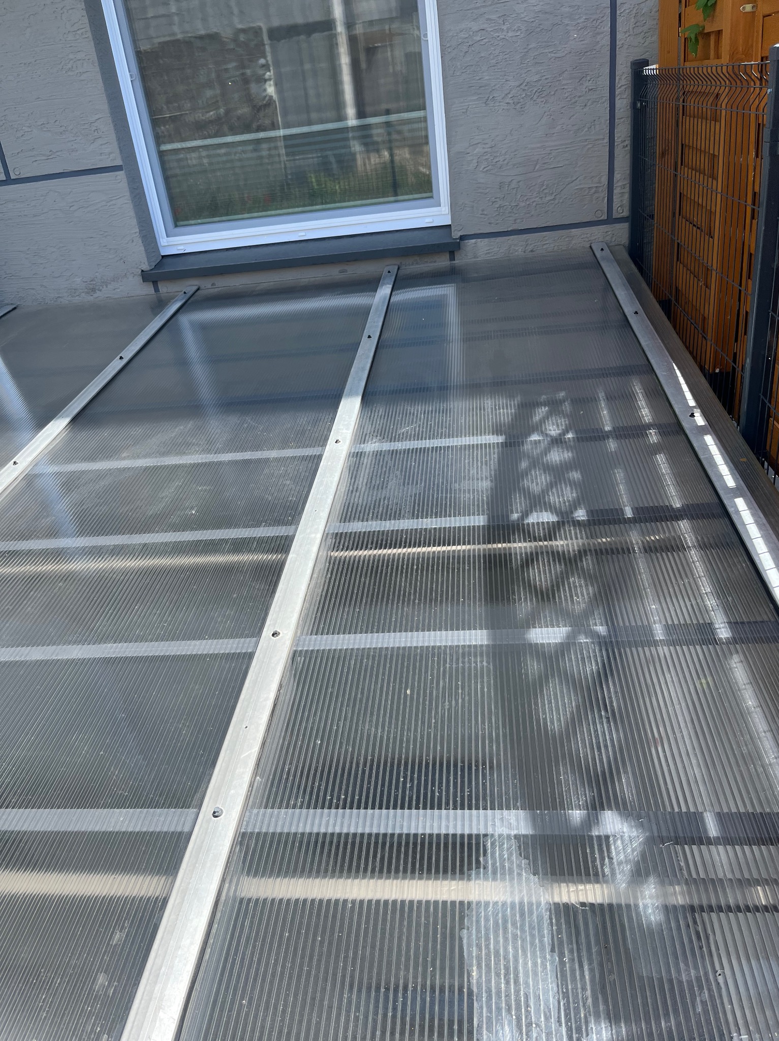Przezroczyste zadaszenie z poliwęglanu, wsparte na metalowej konstrukcji, przylegające do ściany budynku z oknem. Widoczny fragment ogrodzenia.