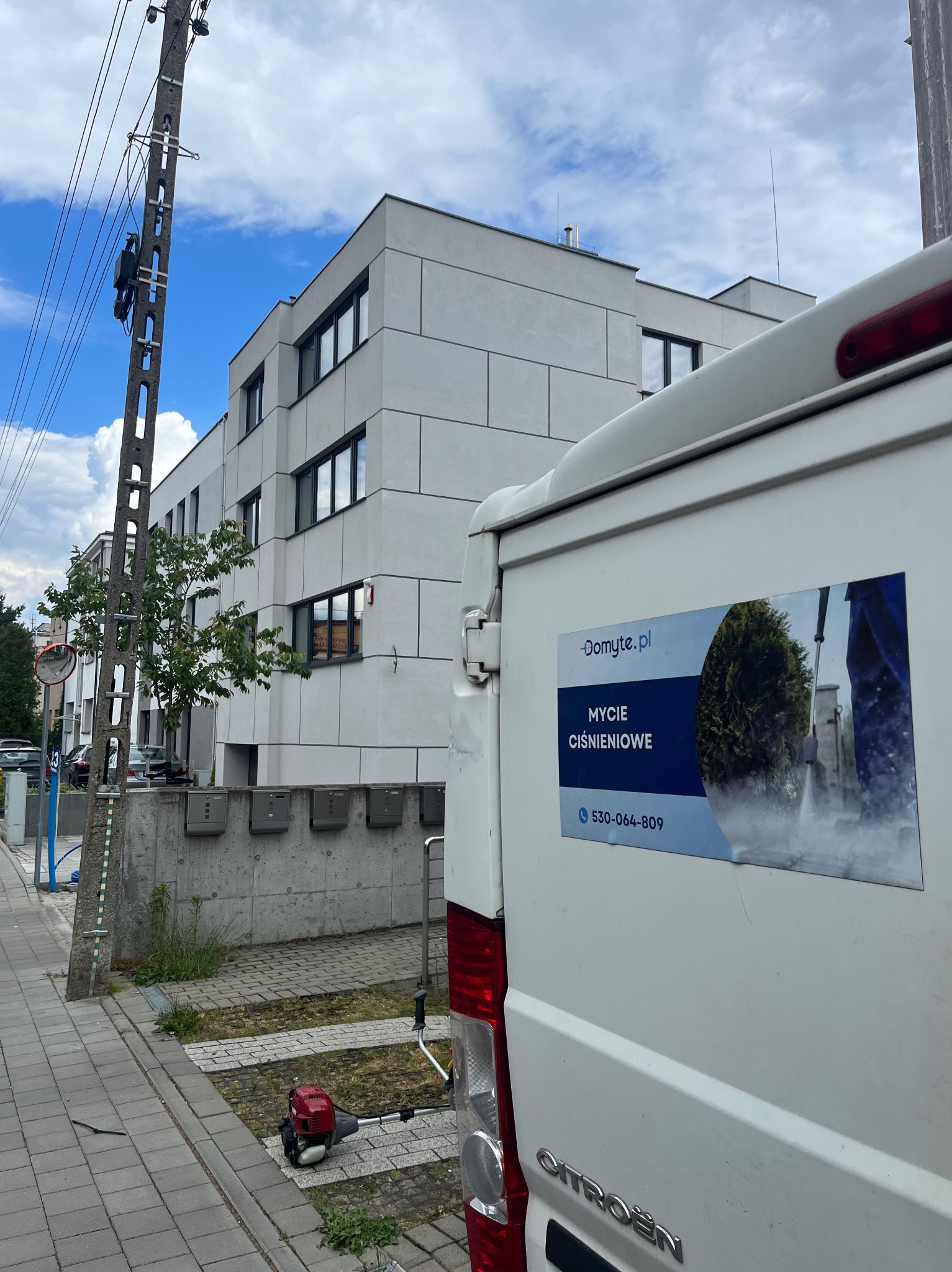 Biały van Citroen z naklejką 'Mycie Ciśnieniowe' na tle nowoczesnego budynku i słupa energetycznego. Obok leży czerwona kosiarka spalinowa.