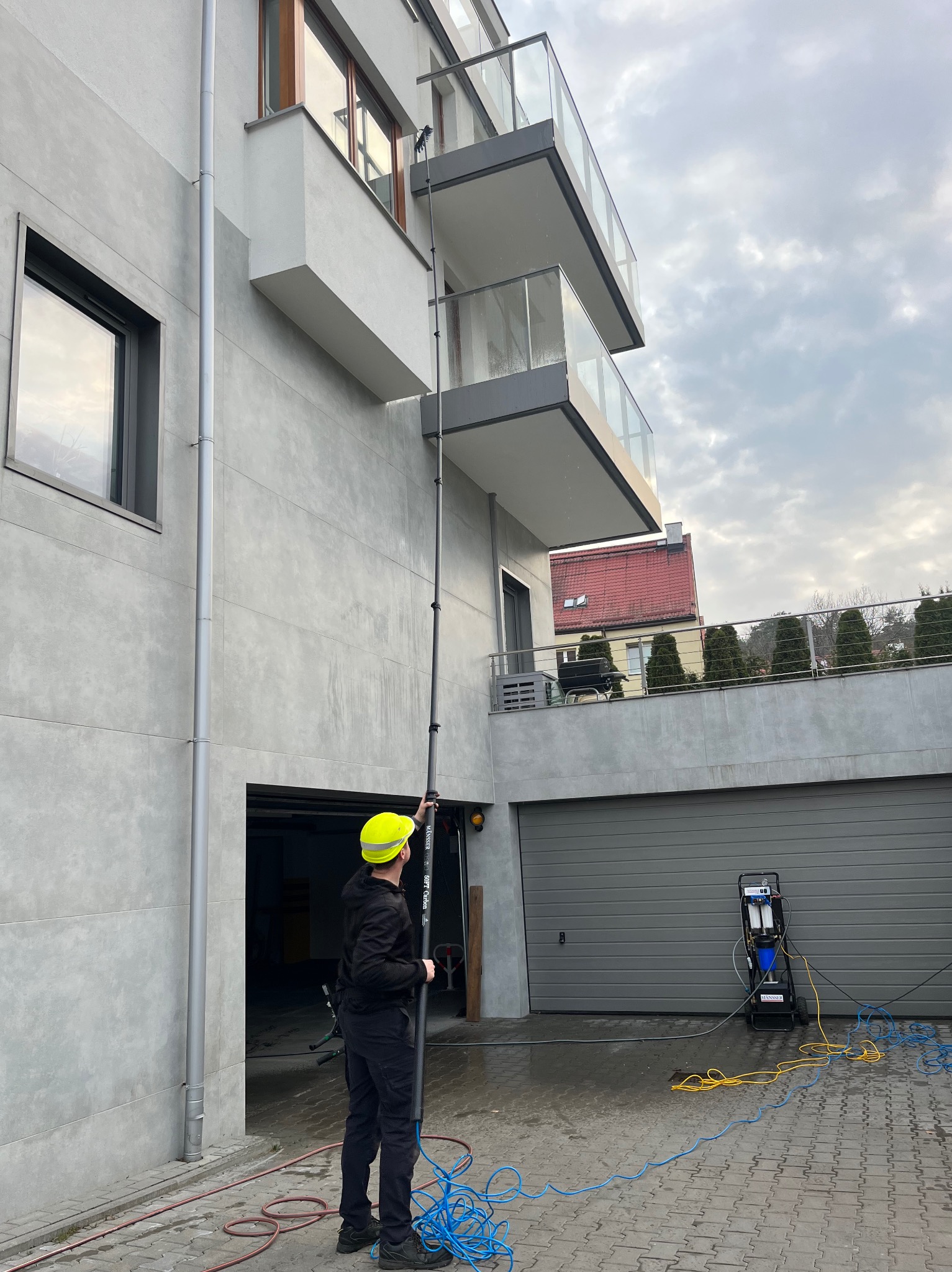 Pracownik w kasku myje elewację domu nowoczesną lancą teleskopową. Widoczny sprzęt do mycia ciśnieniowego i przewody. Szara elewacja, balkony z barierkami.