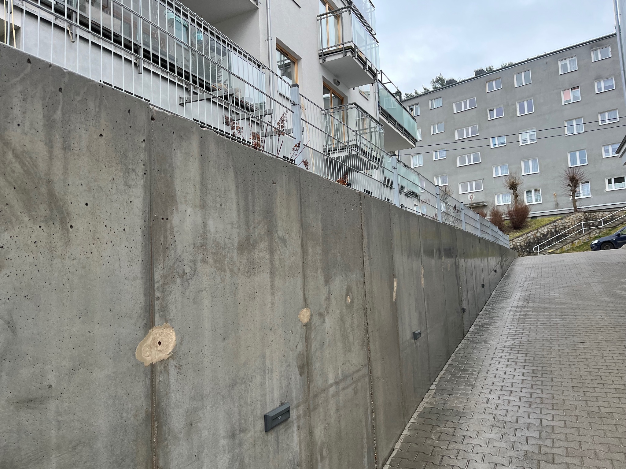 Szara, betonowa ściana oporowa z widocznymi śladami po naprawach, przylegająca do chodnika z kostki brukowej, z budynkiem mieszkalnym w tle. Ujęcie z dołu.