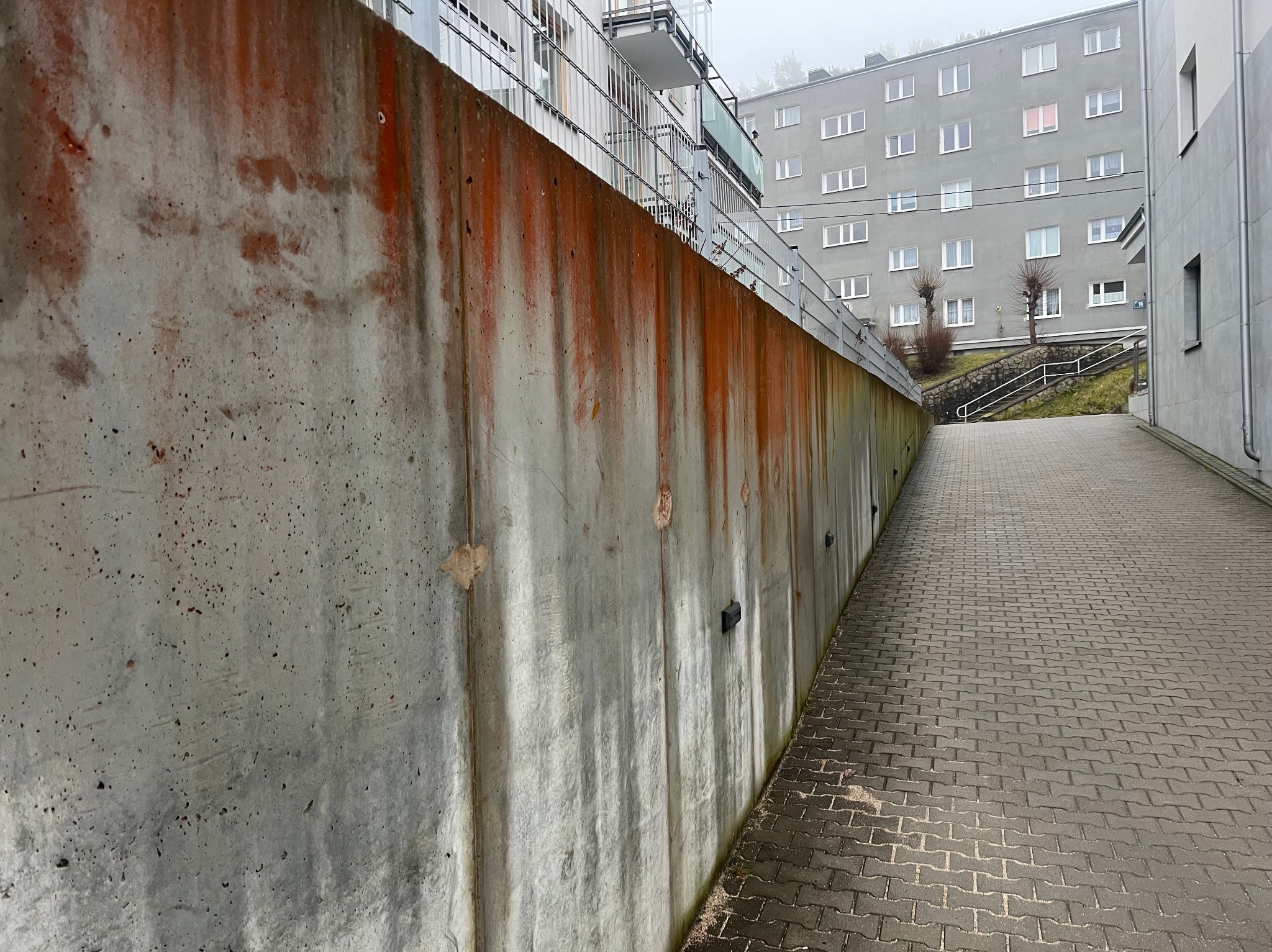 Zewnętrzna ściana budynku z widocznymi zaciekami i zabrudzeniami, obok chodnik z kostki brukowej. W tle budynek mieszkalny z balkonami.