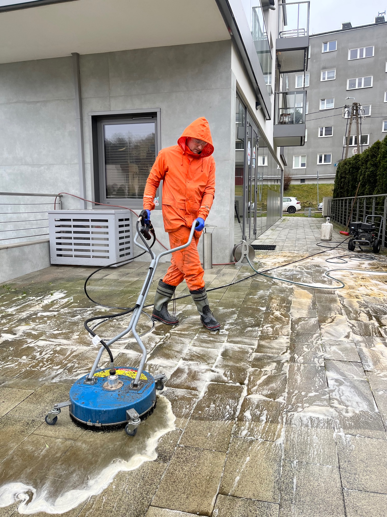 Pracownik w pomarańczowym kombinezonie czyści kostkę brukową na zewnątrz budynku za pomocą specjalistycznej maszyny ciśnieniowej. Widoczne zabrudzenia i piana.