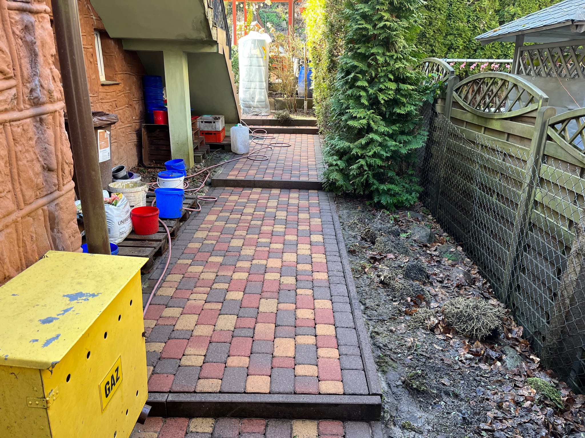 Kolorowa kostka brukowa na schodach prowadzących przez ogród, widoczna skrzynka gazowa i narzędzia, sugerujące prace porządkowe w toku.