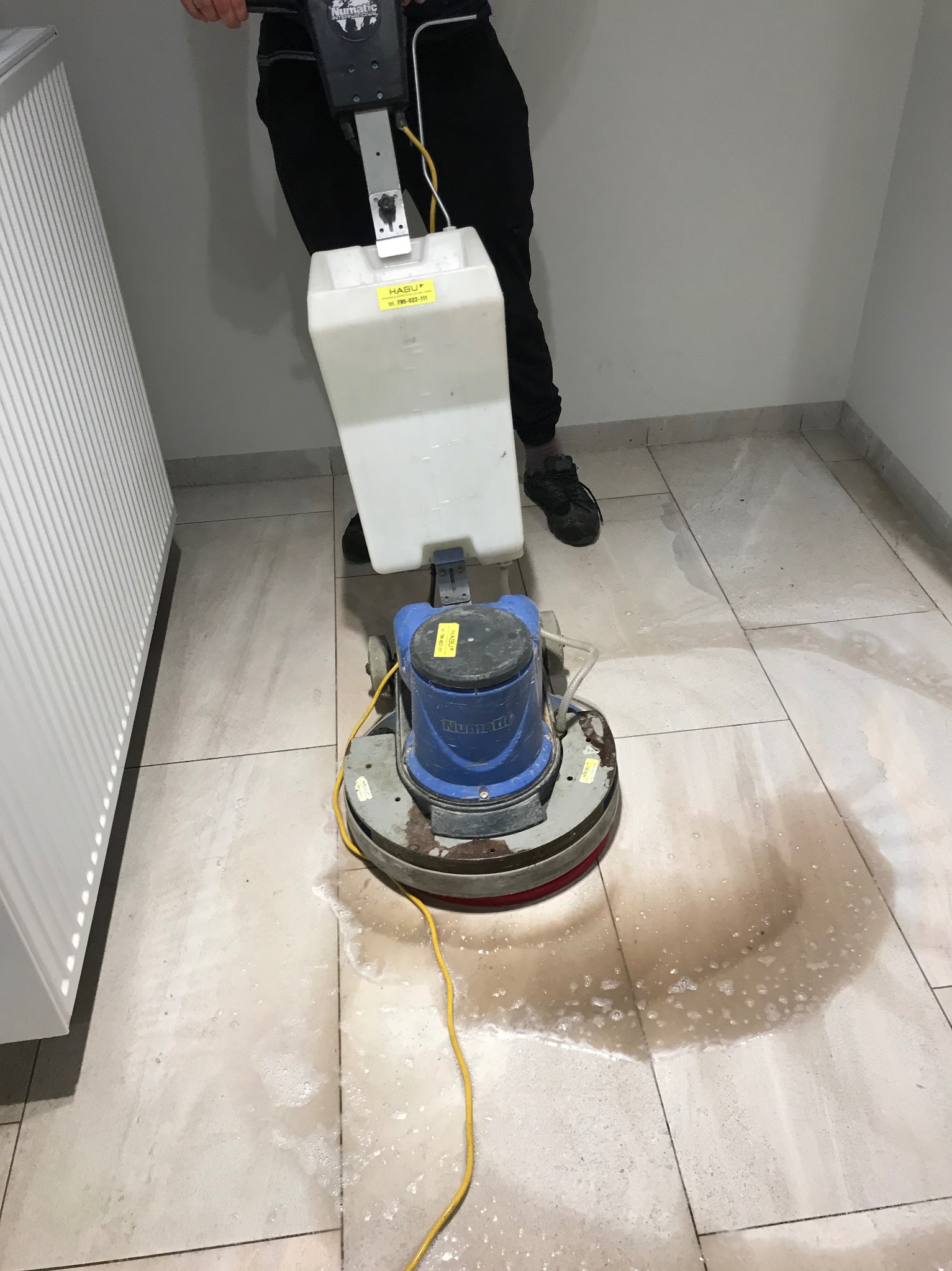 Czyszczenie podłogi z płytek za pomocą profesjonalnej maszyny czyszczącej. Widoczne zabrudzenia i woda na powierzchni. Praca w trakcie wykonywania.