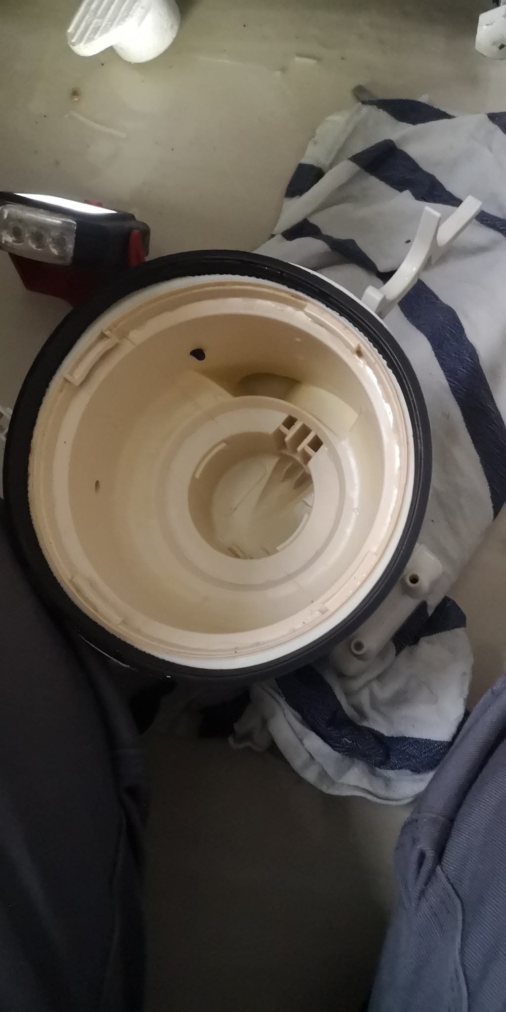 Wnętrze beżowego, okrągłego elementu zmywarki z czarną uszczelką, widoczne otwory i plastikowe wzmocnienia, obok leży szmatka w biało-niebieskie paski i lampa warsztatowa.