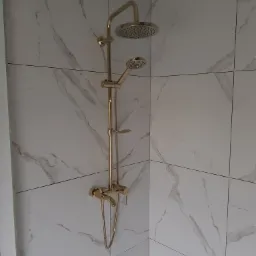 Złota bateria prysznicowa z deszczownicą zamontowana na ścianie wyłożonej białymi płytkami imitującymi marmur, widoczne krzyżyki dystansowe między płytkami.