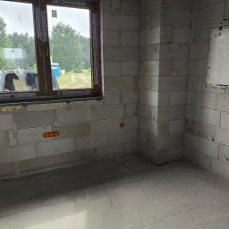 PODMAR MARIUSZ PODLASIŃSKI - Surowy stan wnętrza budynku w trakcie prac wykończeniowych: widoczne murowane ściany z betonu komórkowego, okno z brązową ramą, wystające kable elektryczne w pomarańczowych peszlach oraz otwór...