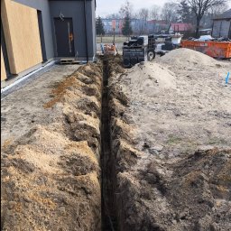 PODMAR MARIUSZ PODLASIŃSKI - Wykop w ziemi z ułożonymi kablami elektrycznymi, widok z góry na tle budowanego domu jednorodzinnego i zgromadzonych materiałów budowlanych.