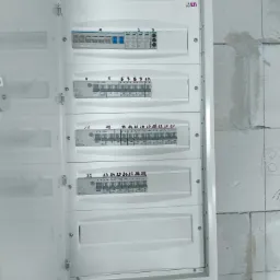 Otwarta, biała skrzynka rozdzielcza z numerowanymi bezpiecznikami na ścianie z betonu komórkowego. Widoczne kable w peszlach. Detal instalacji.