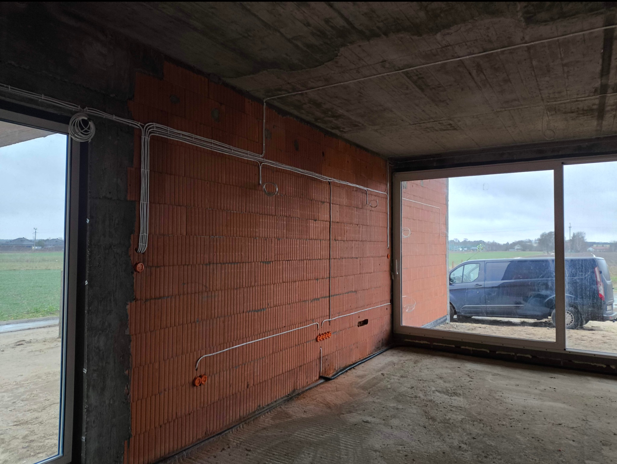 Surowy mur z cegły z widocznym okablowaniem elektrycznym i puszkami instalacyjnymi, w tle zaparkowany ciemny bus.