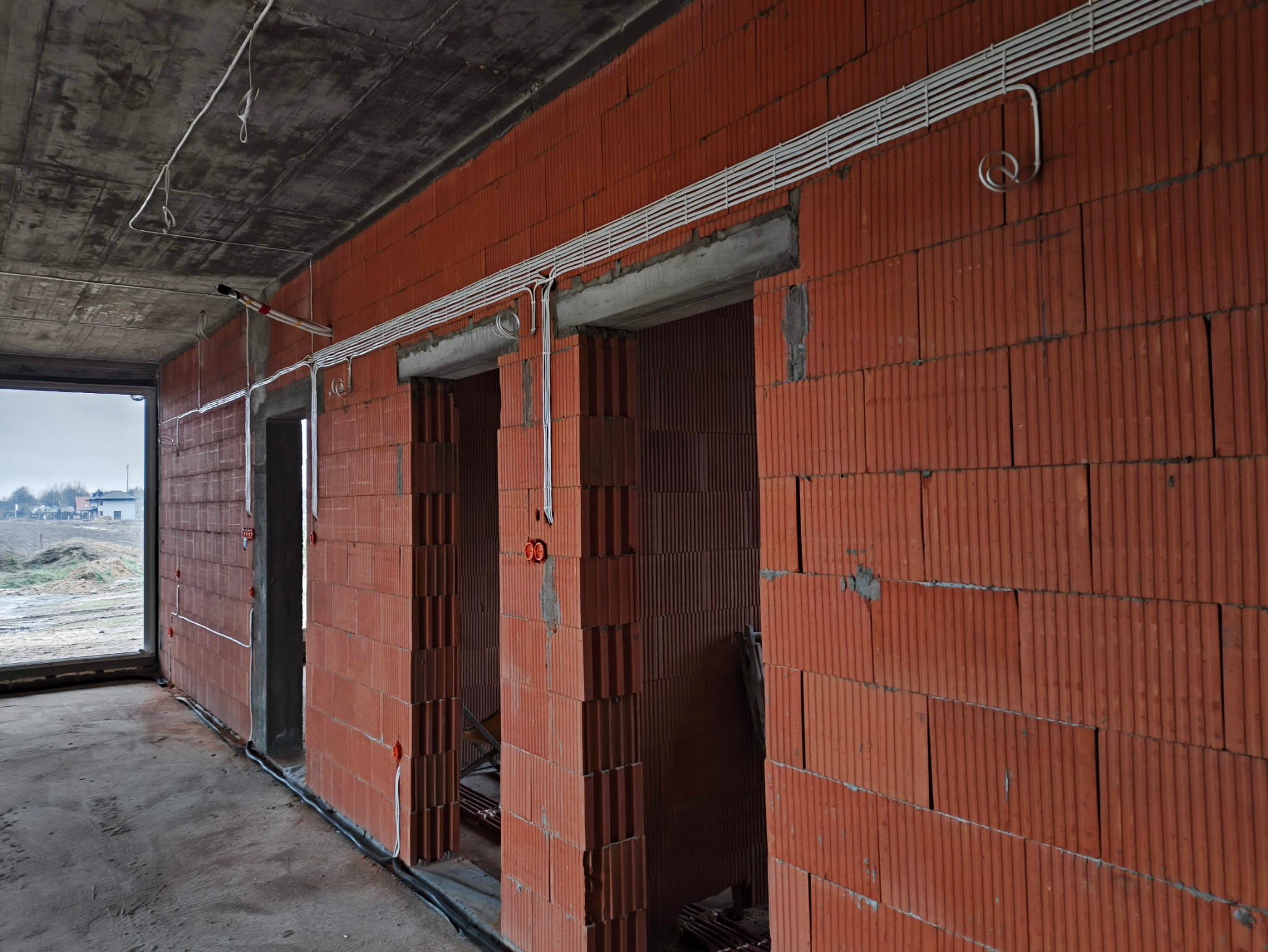 Wewnętrzne ściany budynku w trakcie budowy z widocznymi, poprowadzonymi przewodami elektrycznymi w białych rurkach PCV, zamontowanymi gniazdami i puszkami elektrycznymi. Surowy betonowy strop...