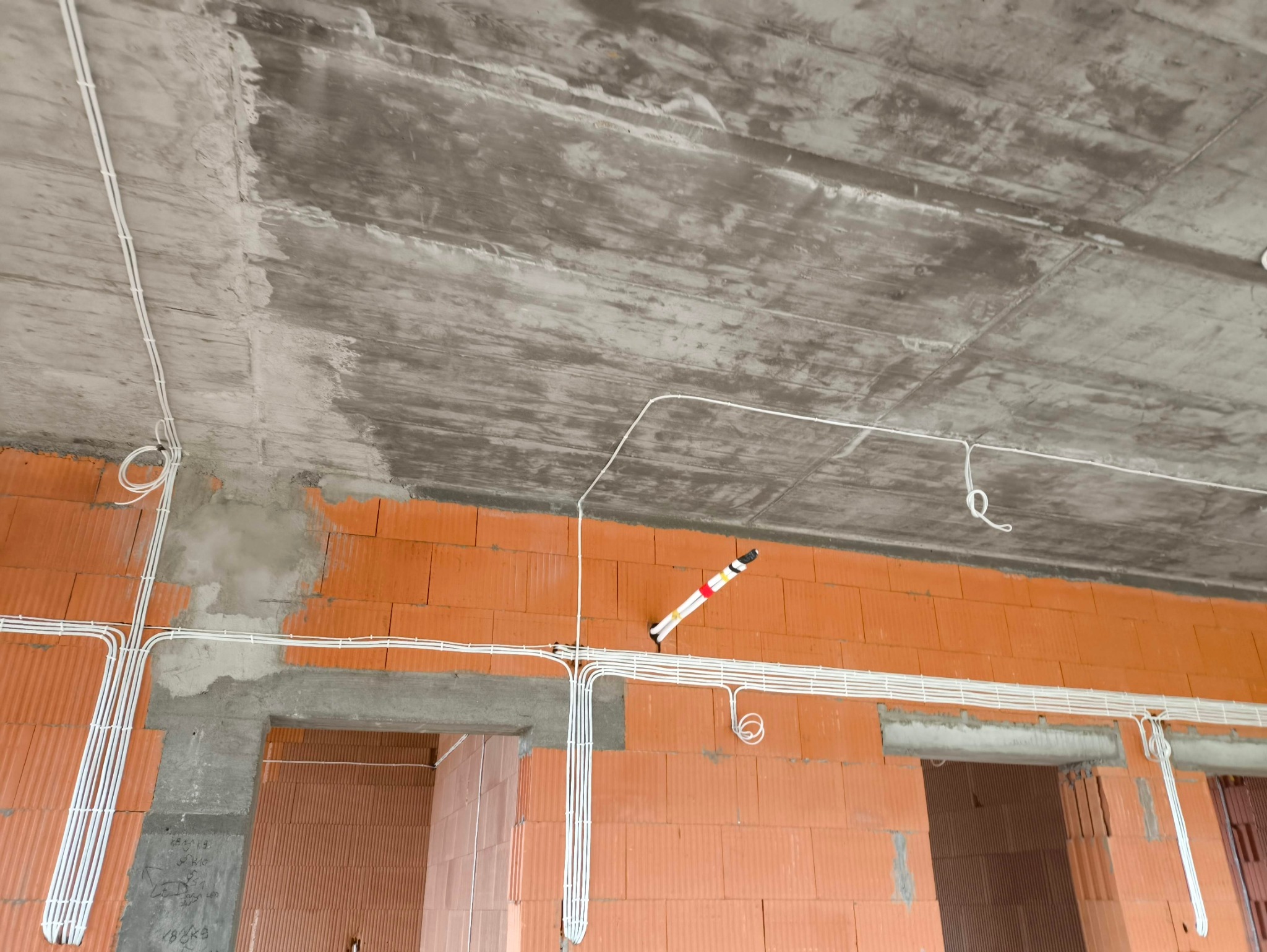 Białe kable elektryczne ułożone na surowej ścianie z czerwonej cegły i betonowym suficie w trakcie budowy domu, widoczne otwory drzwiowe.