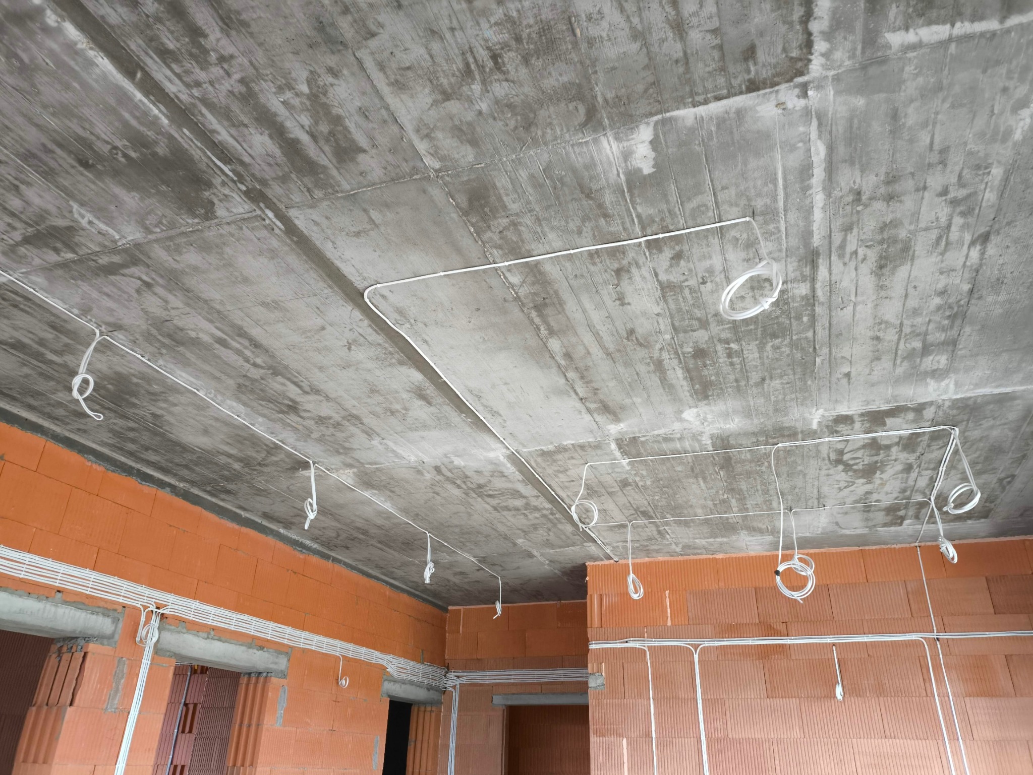 Surowy betonowy strop z widocznymi przewodami elektrycznymi, pętle kabli zwisają, ściany z czerwonej cegły.