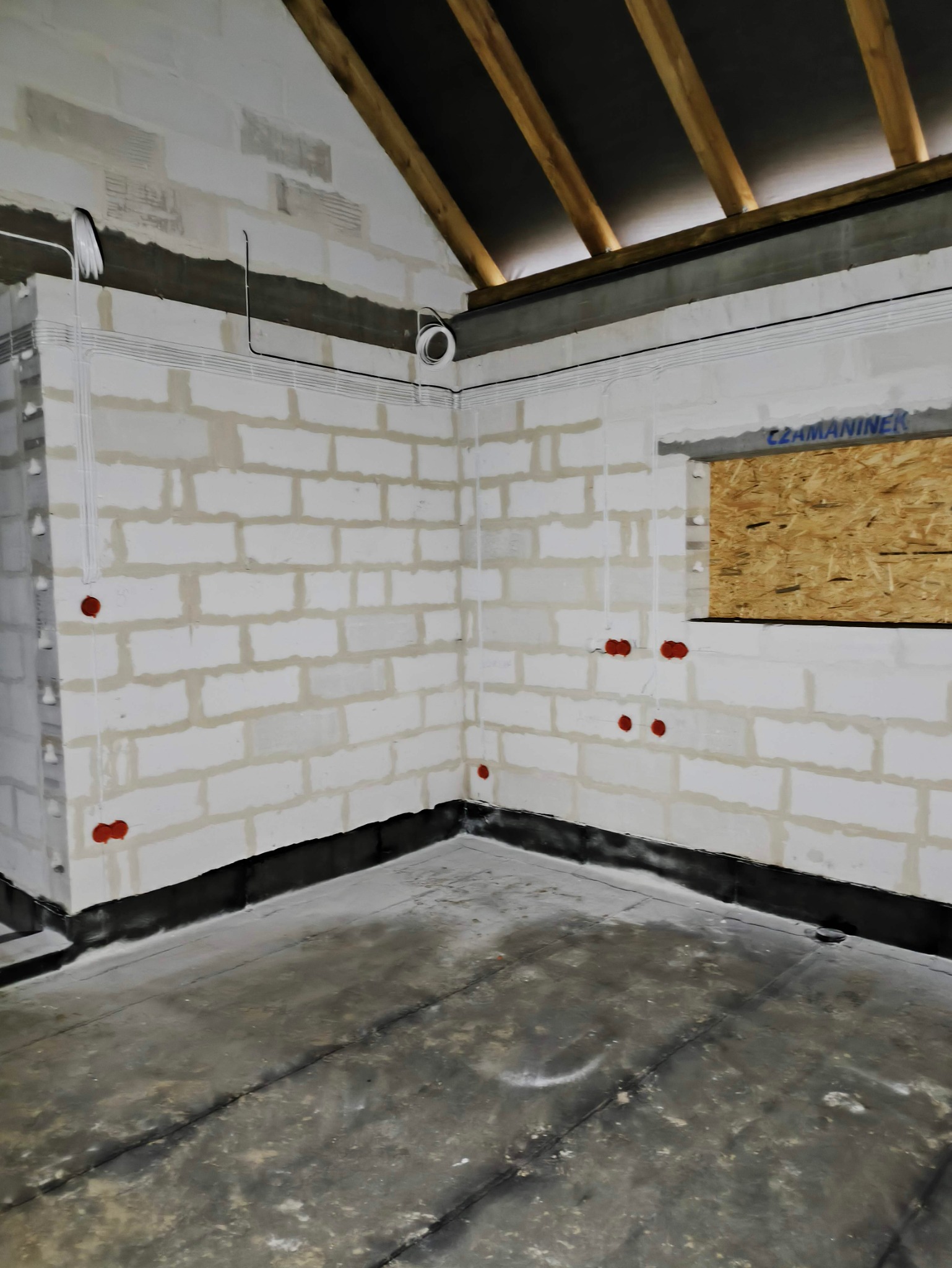 Surowy stan wnętrza w trakcie budowy, widoczne rozprowadzone przewody elektryczne w białych rurkach na ścianach z betonu komórkowego, przygotowane punkty pod gniazdka w kolorze czerwonym oraz sufit...