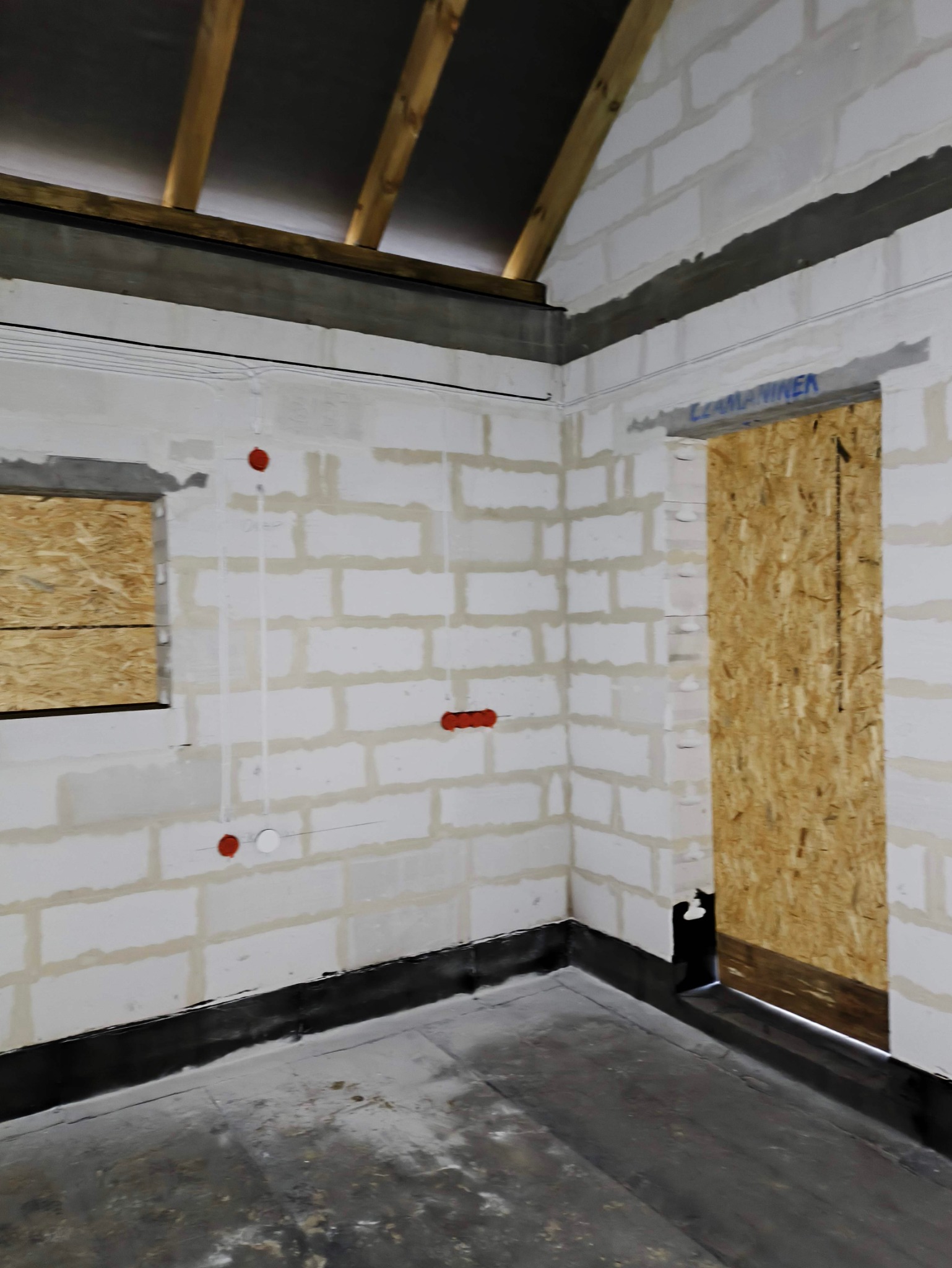 Wnętrze w trakcie prac wykończeniowych: ściany z białych bloczków z widocznymi przewodami elektrycznymi oznaczonymi czerwonymi puszkami, okno i otwór drzwiowy zabite płytą OSB, surowy beton...