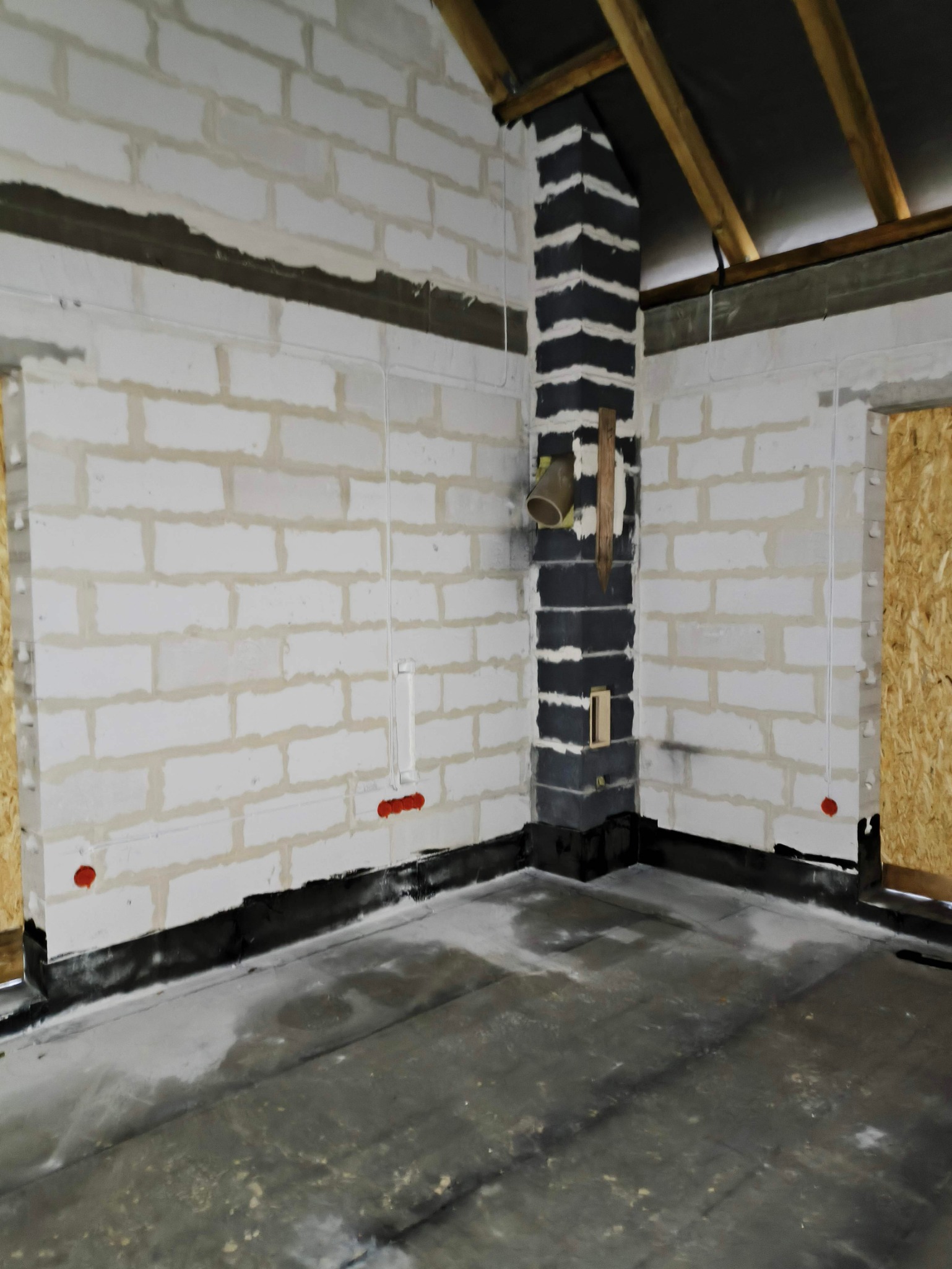 Surowy stan wnętrza budynku w trakcie prac: widoczne białe ściany z betonu komórkowego, czarna izolacja przeciwwilgociowa przy podłodze, pionowe bruzdy pod przewody elektryczne oraz komin...