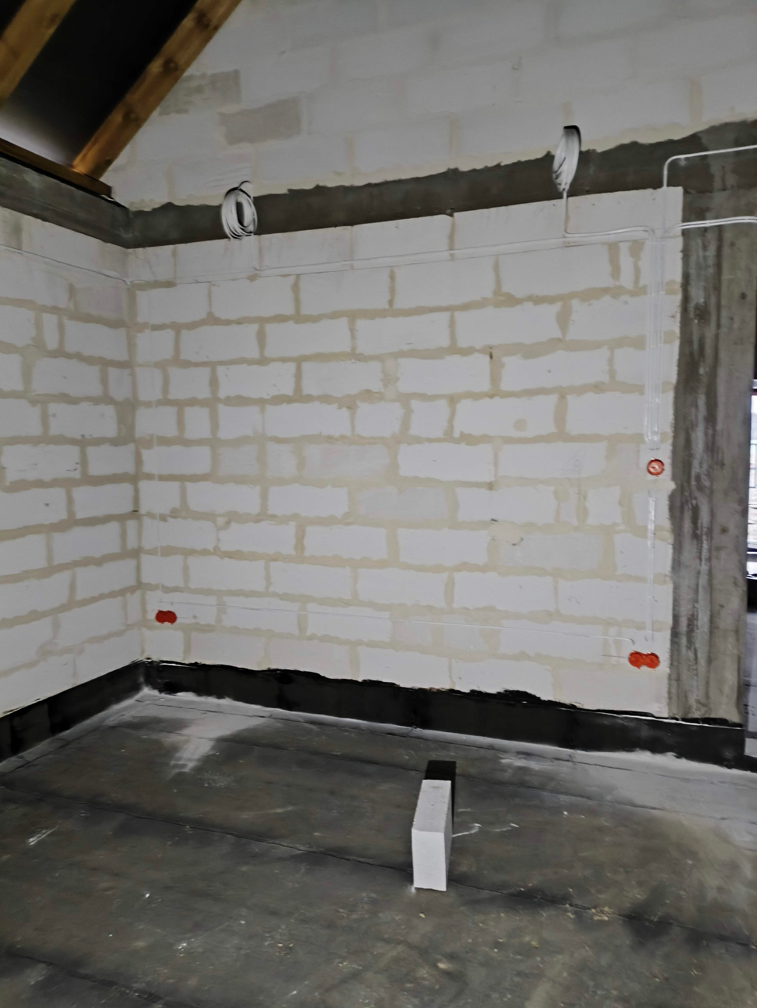 Wnętrze w trakcie instalacji elektrycznej: ściany z betonu komórkowego, widoczne przewody i puszki instalacyjne, surowy beton na podłodze, fragment więźby dachowej