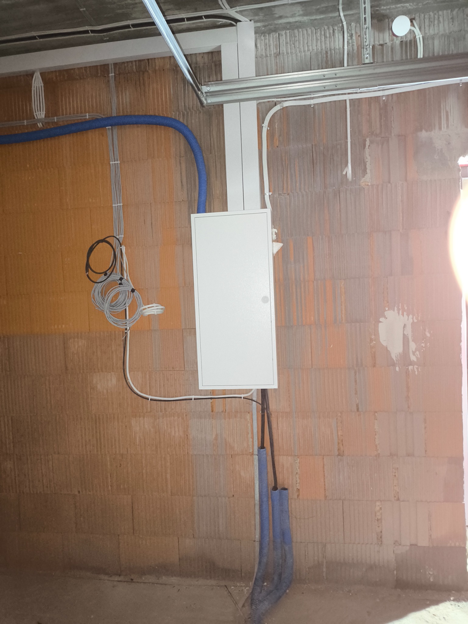 Biała skrzynka elektryczna zamontowana na ścianie z cegły, z niebieskimi rurami karbowanymi i przewodami, widoczne elementy konstrukcji sufitu.