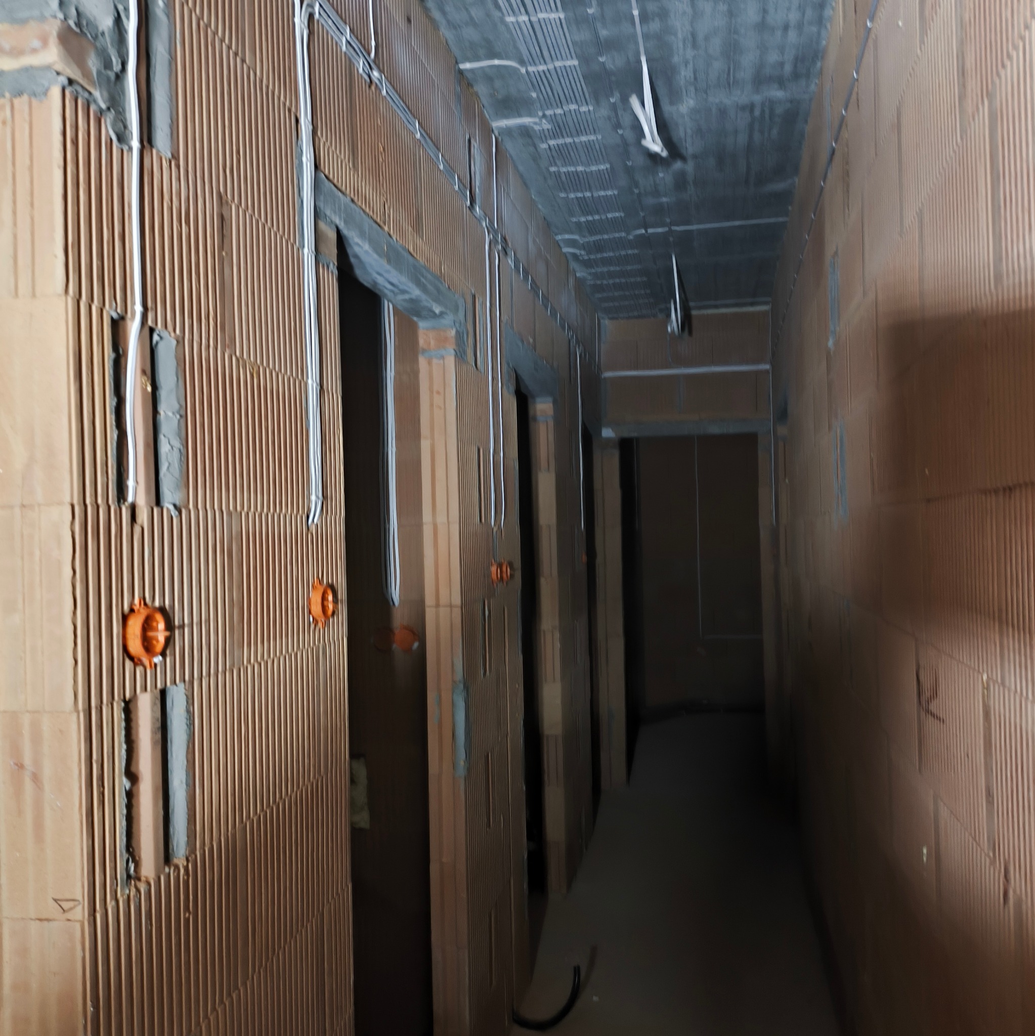 Korytarz w surowym stanie z instalacją elektryczną: widoczne kable prowadzone po ścianach z cegły ceramicznej oraz puszki instalacyjne w kolorze pomarańczowym.