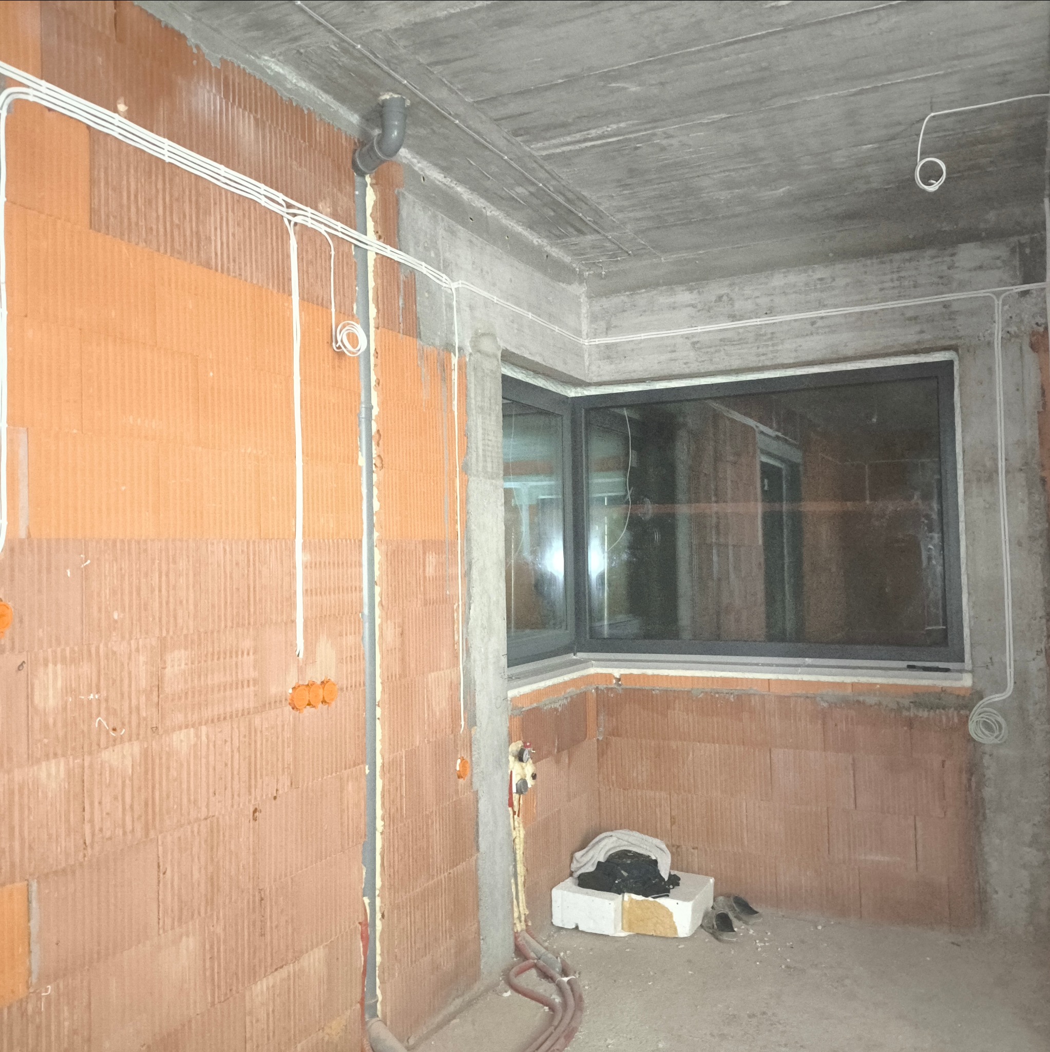 Surowy stan: instalacja elektryczna w budynku z cegły, widoczne kable prowadzone po ścianach i suficie, puszki elektryczne w ścianach, okno w szarej ramie, betonowy sufit.