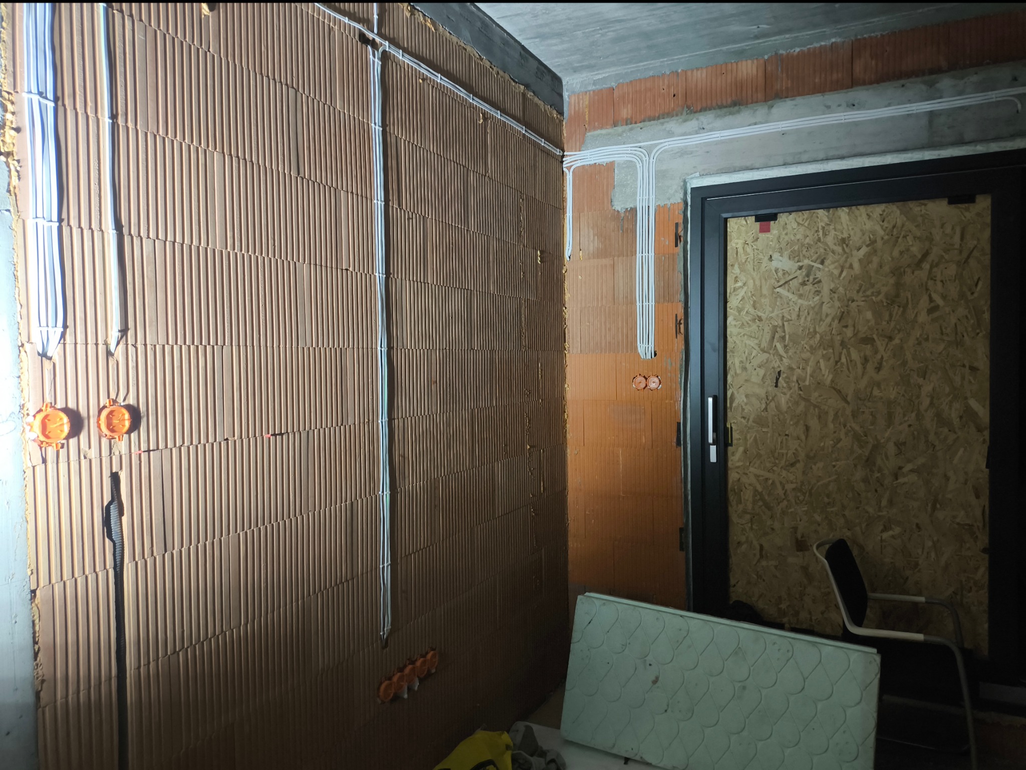 Wnętrze w trakcie prac elektrycznych: widoczne kable prowadzone po ścianach z pustaków ceramicznych, puszki instalacyjne i zabezpieczone drzwi wejściowe.