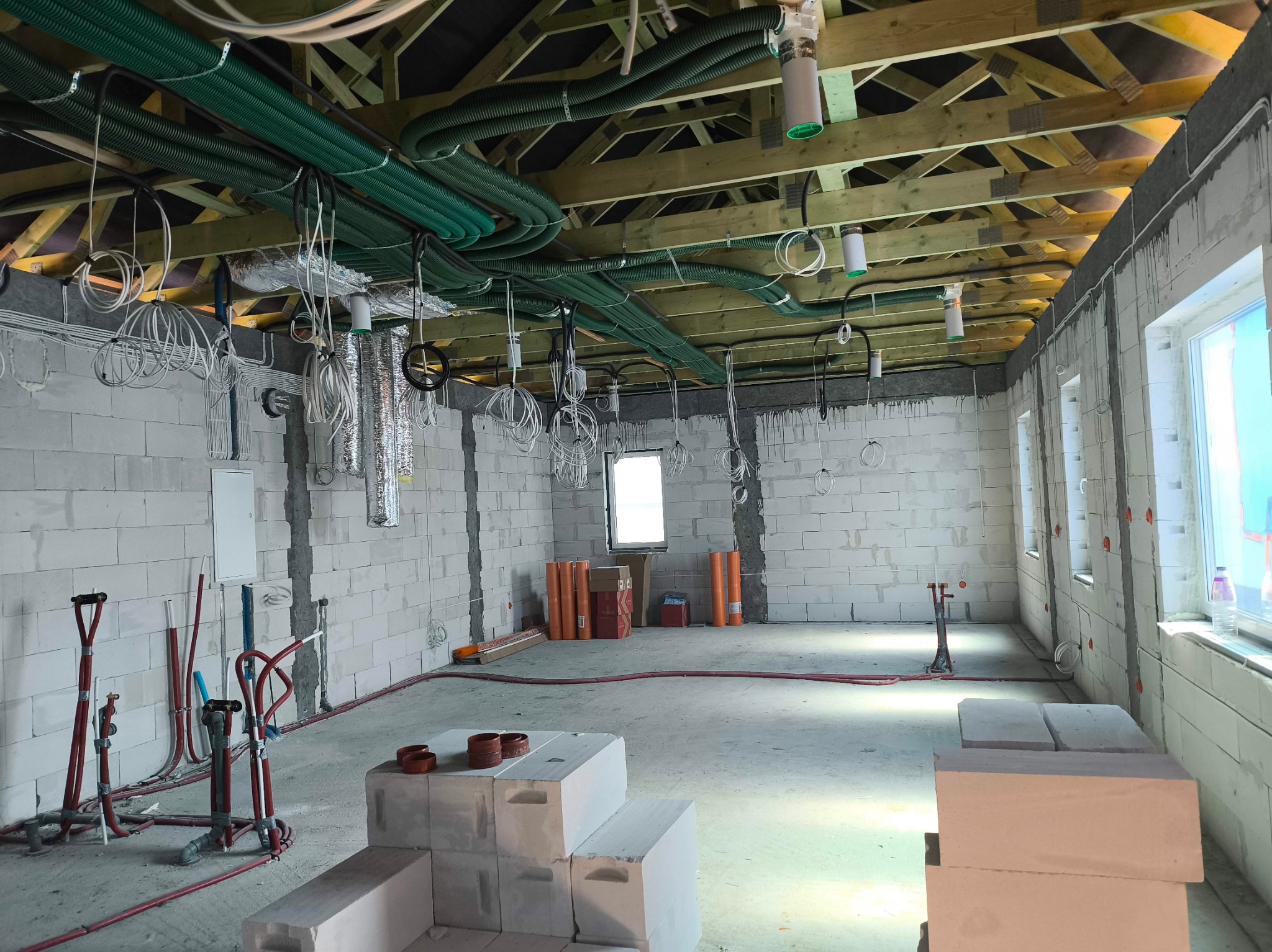Surowy stan: widok wnętrza z instalacjami pod stropem, rury wentylacyjne i kable elektryczne zwisają. Białe ściany z betonu komórkowego, okna w tle.