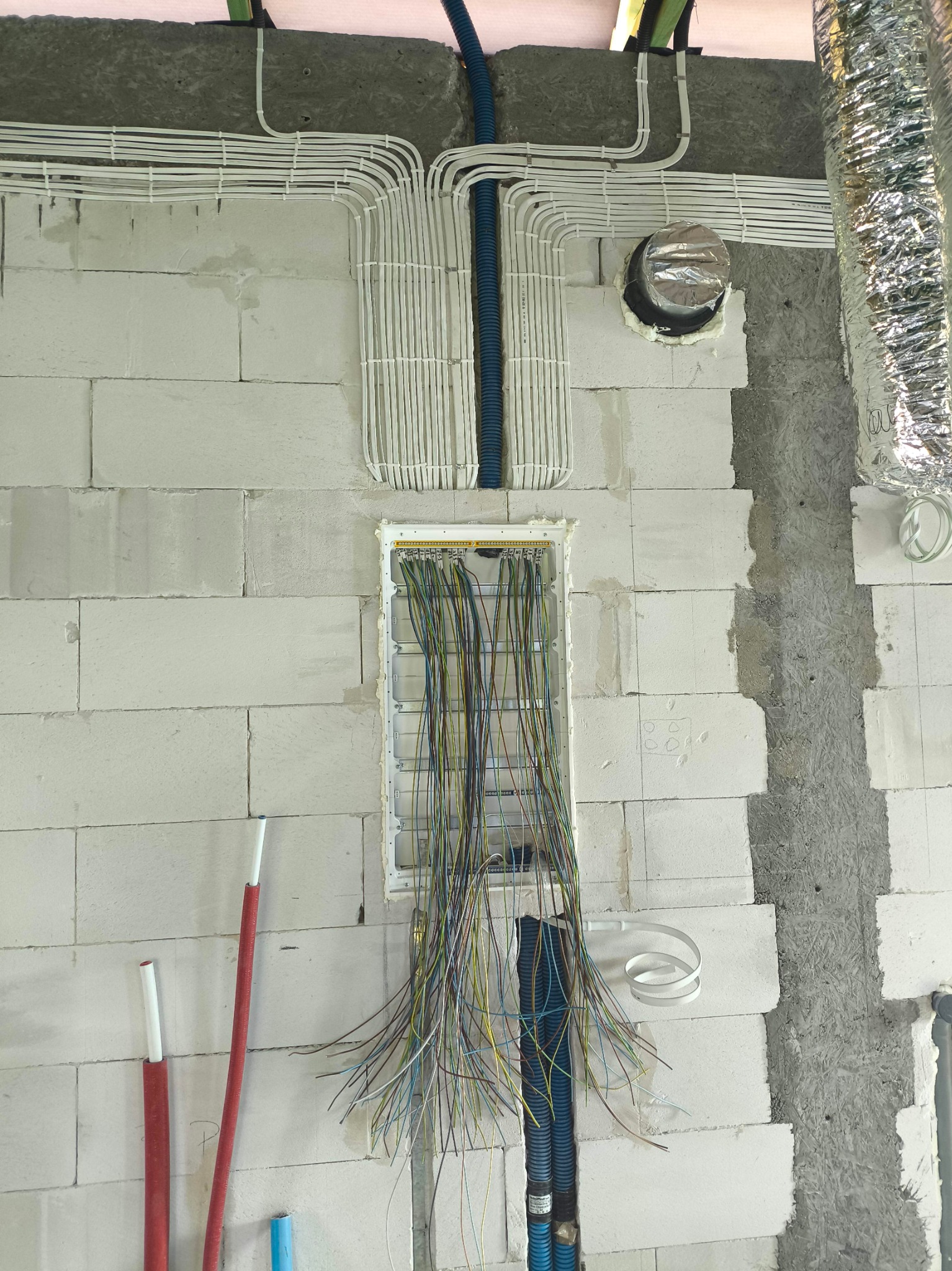 Rozprowadzenie przewodów elektrycznych w białej ścianie z betonu komórkowego, widoczne kable w peszlach i puszce. Elementy instalacji wentylacyjnej w tle.