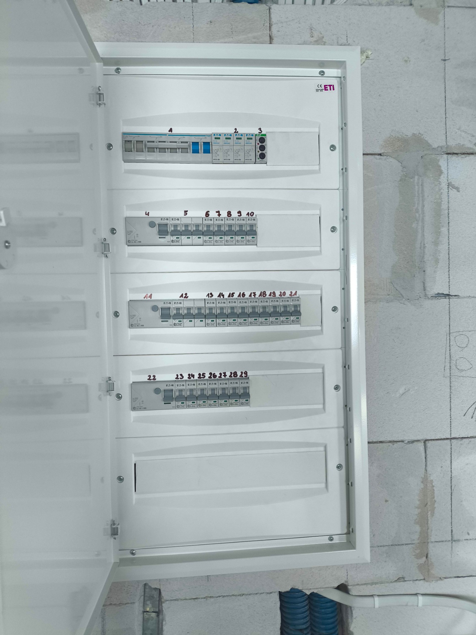 Otwarta, biała skrzynka rozdzielcza z numerowanymi bezpiecznikami na ścianie z betonu komórkowego. Widoczne kable w peszlach. Detal instalacji.