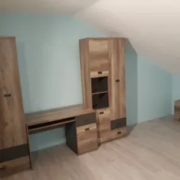 Zestaw mebli w pokoju na poddaszu: szafa, biurko i komoda w odcieniach drewna z ciemnoszarymi akcentami, na jasnej podłodze, ściany w kolorze błękitnym.