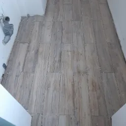 Układanie drewnopodobnych płytek podłogowych w łazience, widoczne krzyżyki dystansowe, surowy stan wykończenia ścian i rur kanalizacyjnych.