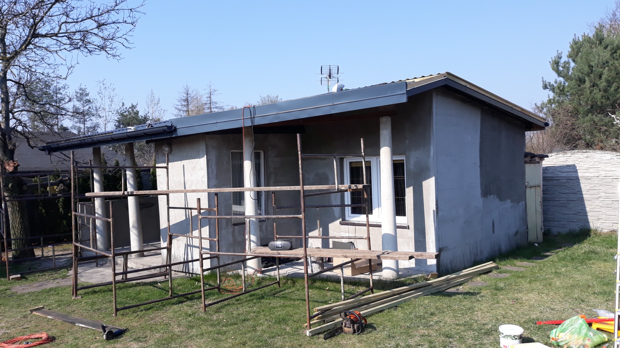 Dom w trakcie prac elewacyjnych, widoczne rusztowania, świeżo otynkowana ściana, przygotowane deski i piła łańcuchowa na trawie.