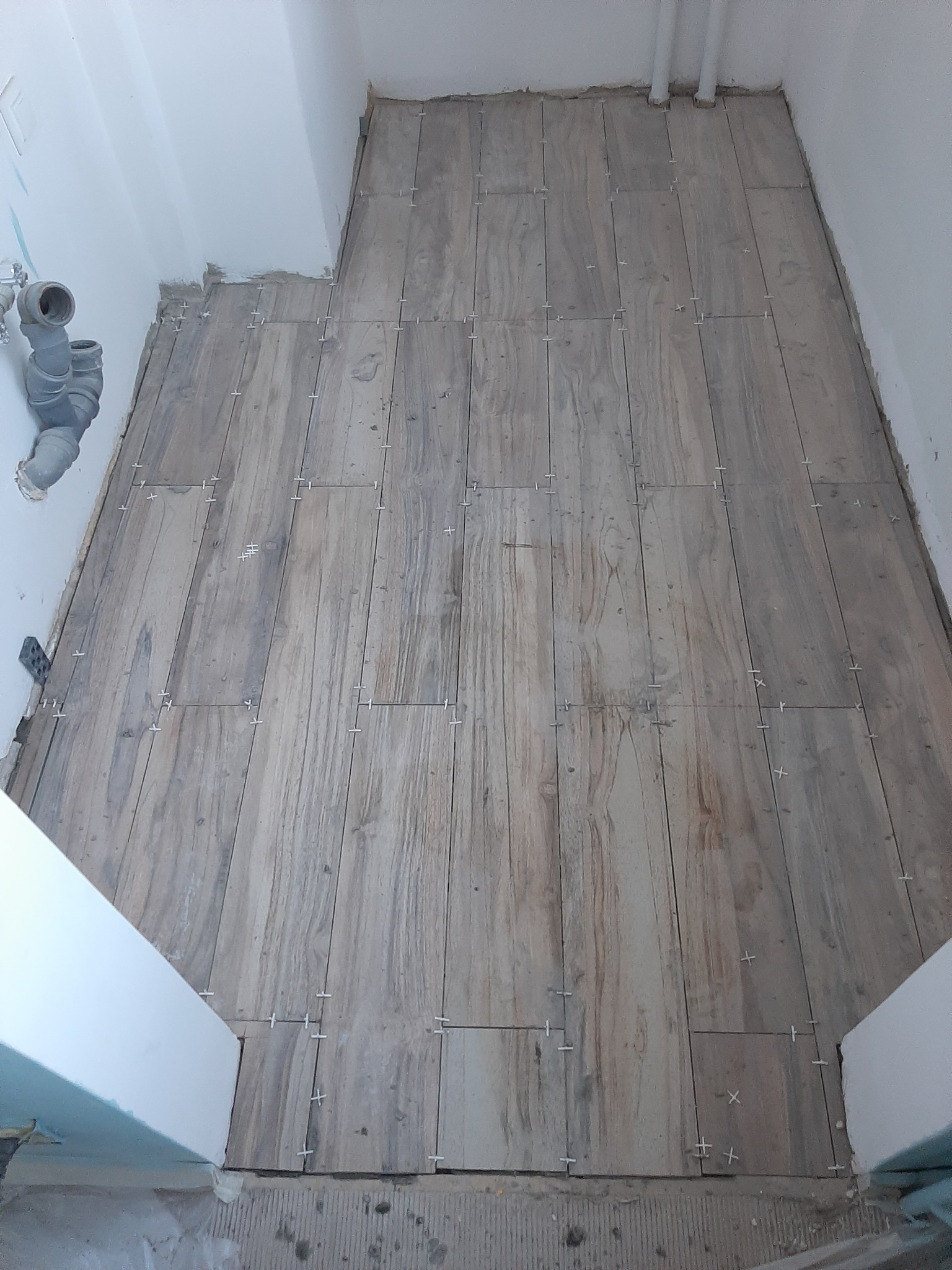 Układanie drewnopodobnych płytek podłogowych w łazience, widoczne krzyżyki dystansowe, surowy stan wykończenia ścian i rur kanalizacyjnych.