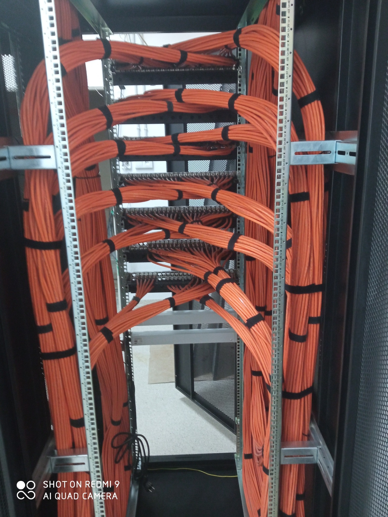 Pomarańczowe kable telekomunikacyjne uporządkowane w szafie rack, spięte czarnymi opaskami, widoczne metalowe szyny montażowe z numeracją jednostek U