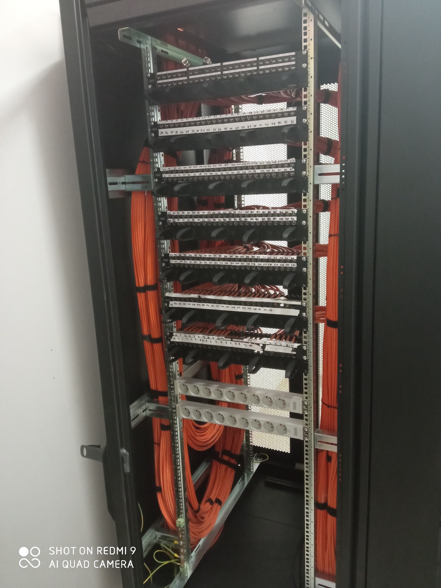 Rzut z dołu na szafę rackową z uporządkowanymi pomarańczowymi kablami sieciowymi, patch panele z opisami portów oraz listwa zasilająca z gniazdkami Schuko.
