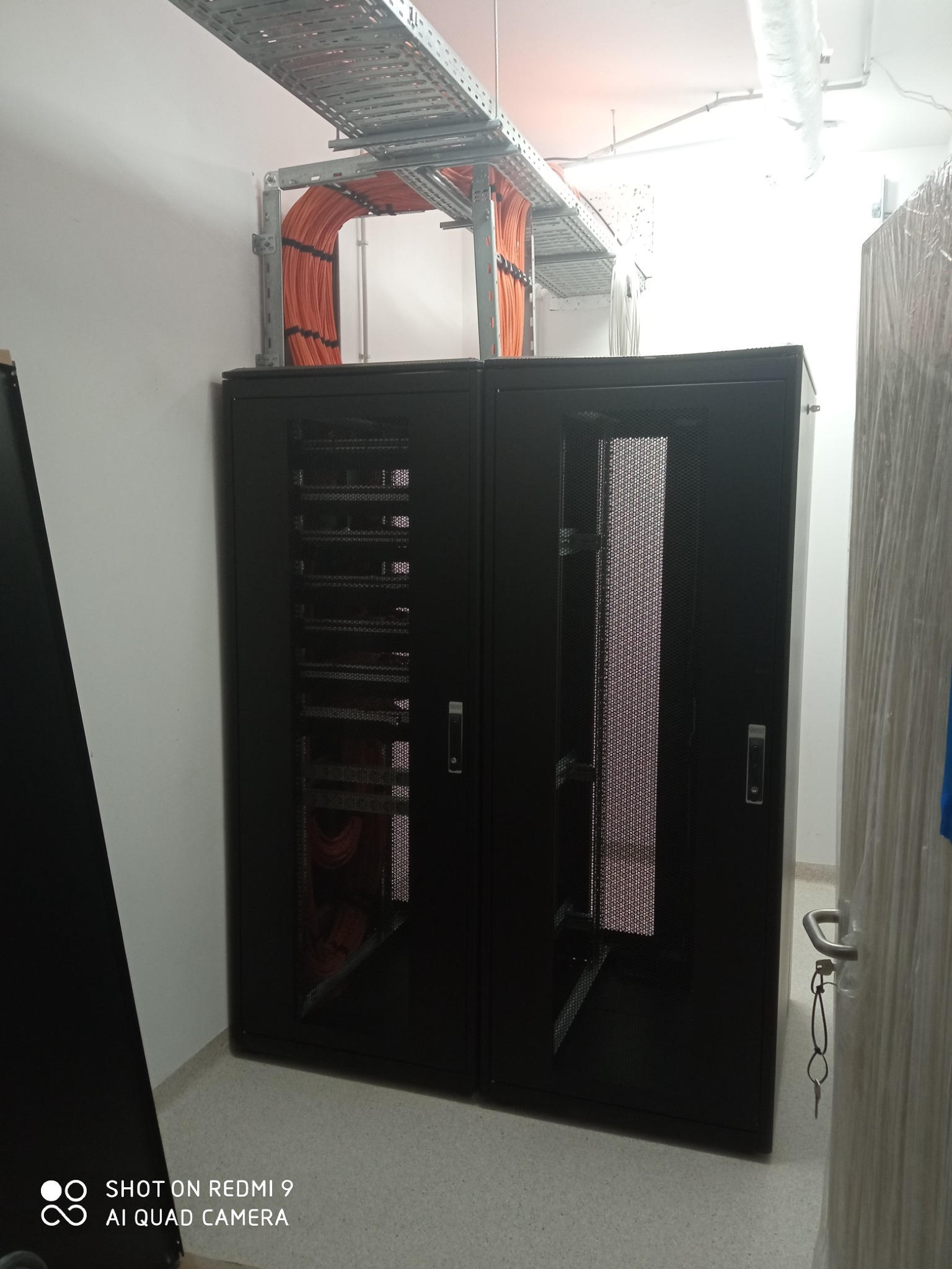 Dwie szafy serwerowe w pomieszczeniu technicznym z korytem kablowym pod sufitem wypełnionym pomarańczowymi kablami.