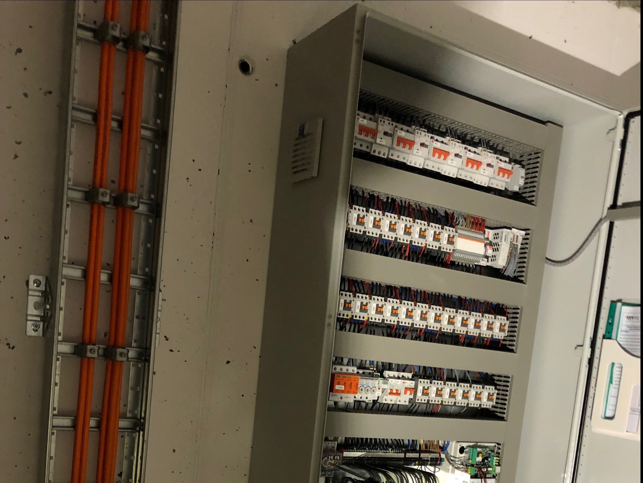 Otwarta szafa sterownicza z widocznymi bezpiecznikami, przekaźnikami i okablowaniem, obok pionowy kanał z pomarańczowymi przewodami elektrycznymi przymocowanymi metalowymi uchwytami do ściany.