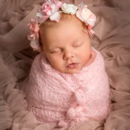 Śpiący noworodek otulony różowym kocykiem, z wiankiem z różowych i białych kwiatów na głowie, ujęcie z góry na tle beżowej tkaniny, Bełchatów.