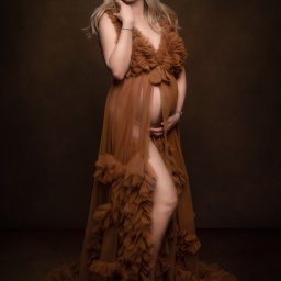 Paulina Pawelus Fotografia - Kobieta w ciąży, ubrana w zwiewną, brązową suknię z falbanami, pozuje na ciemnobrązowym tle, delikatnie dotykając szyi i trzymając dłonie na brzuchu.