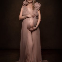 Paulina Pawelus Fotografia - Kobieta w ciąży, w zwiewnej, pudrowo-różowej sukni z bufkami na ramionach, pozuje na ciemnobrązowym tle, dłonie ułożone na brzuchu, delikatny uśmiech.