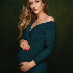 Paulina Pawelus Fotografia - Portret kobiety w ciąży w zielonym body na tle teksturowanej zieleni, z dłońmi splecionymi na brzuchu, delikatna biżuteria i manicure w kolorze neonowego różu.
