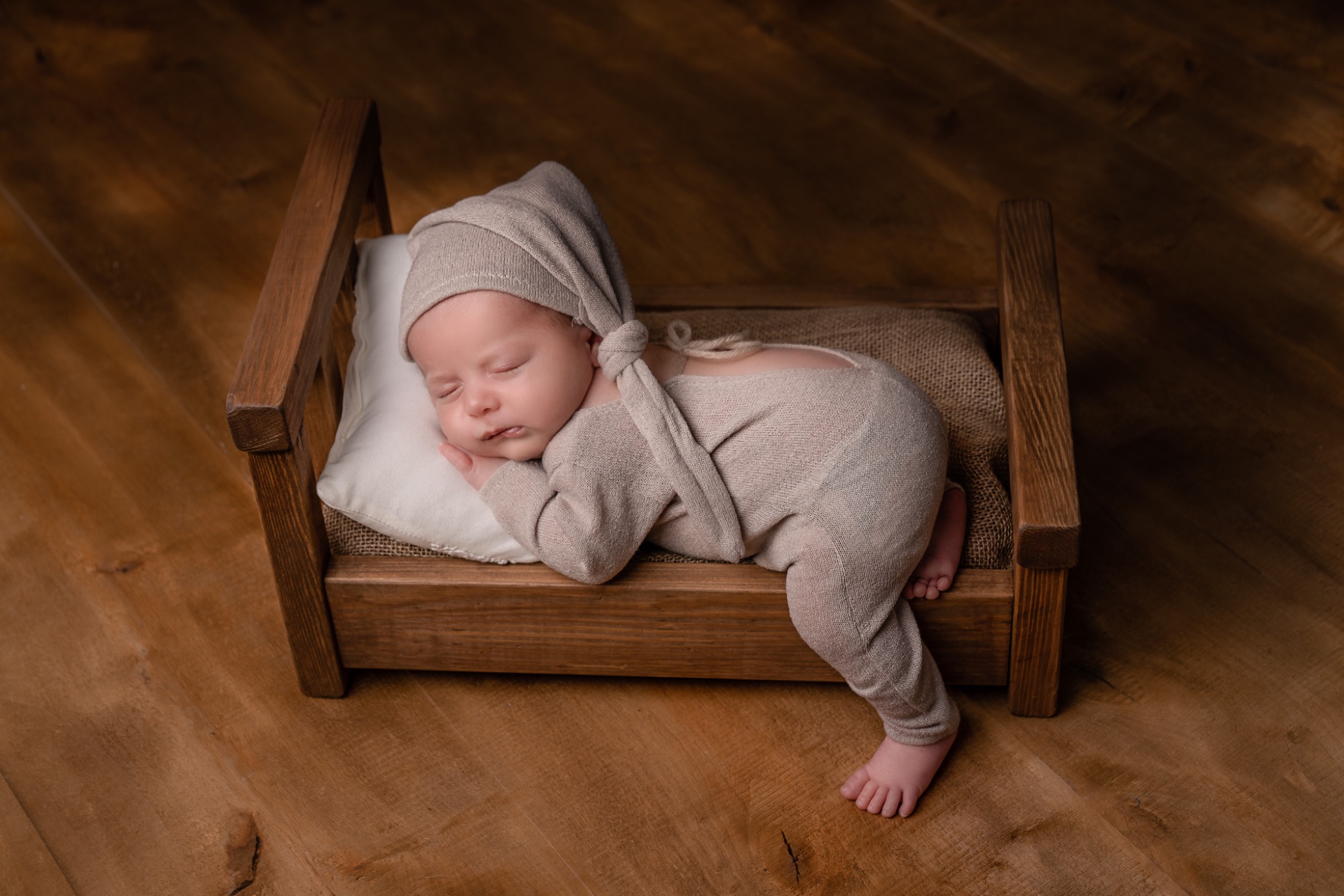 Śpiący noworodek w beżowym stroju i czapce z supełkiem, ułożony na małym, drewnianym łóżeczku z białą poduszką, na drewnianej podłodze.
