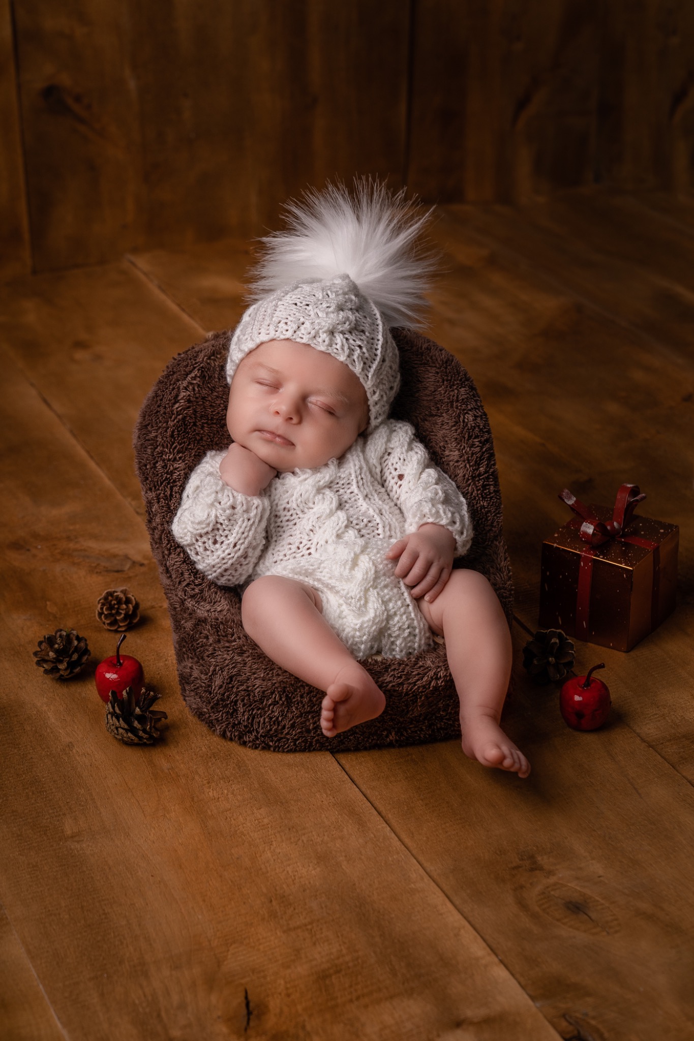 Śpiące niemowlę w białym, dzierganym kombinezonie i czapce z pomponem, ułożone w brązowym, futerkowym gnieździe na drewnianej podłodze, obok jabłka, szyszek i małego prezentu.