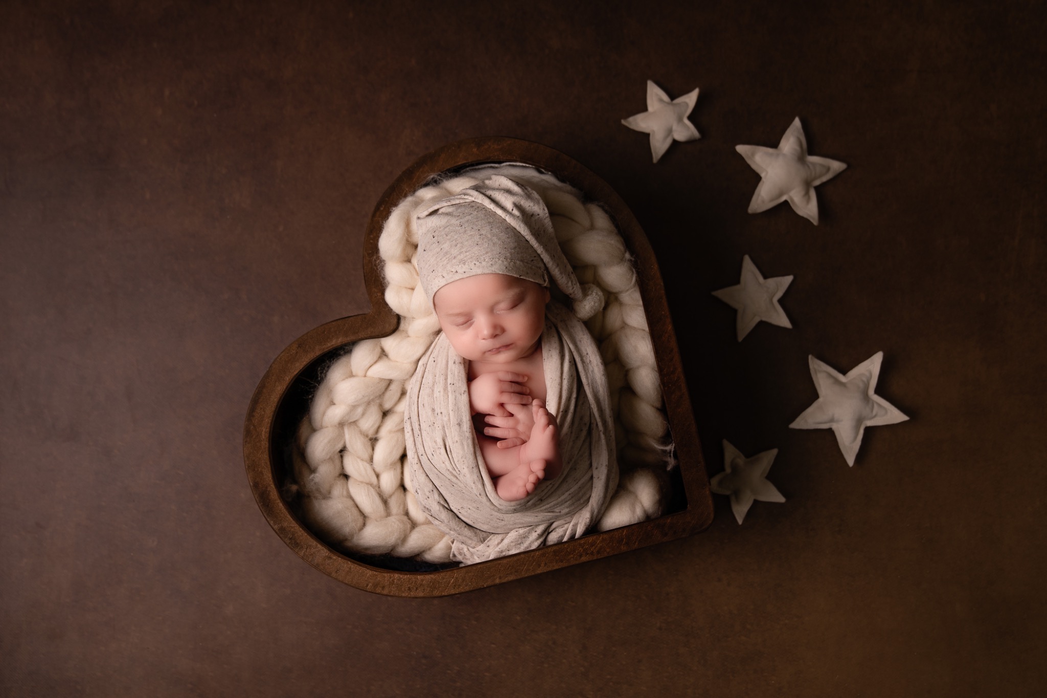 Śpiące nowonarodzone dziecko w beżowym otulaczu i czapeczce, ułożone w drewnianym sercu wypełnionym wełną, otoczone pluszowymi gwiazdkami na brązowym tle.