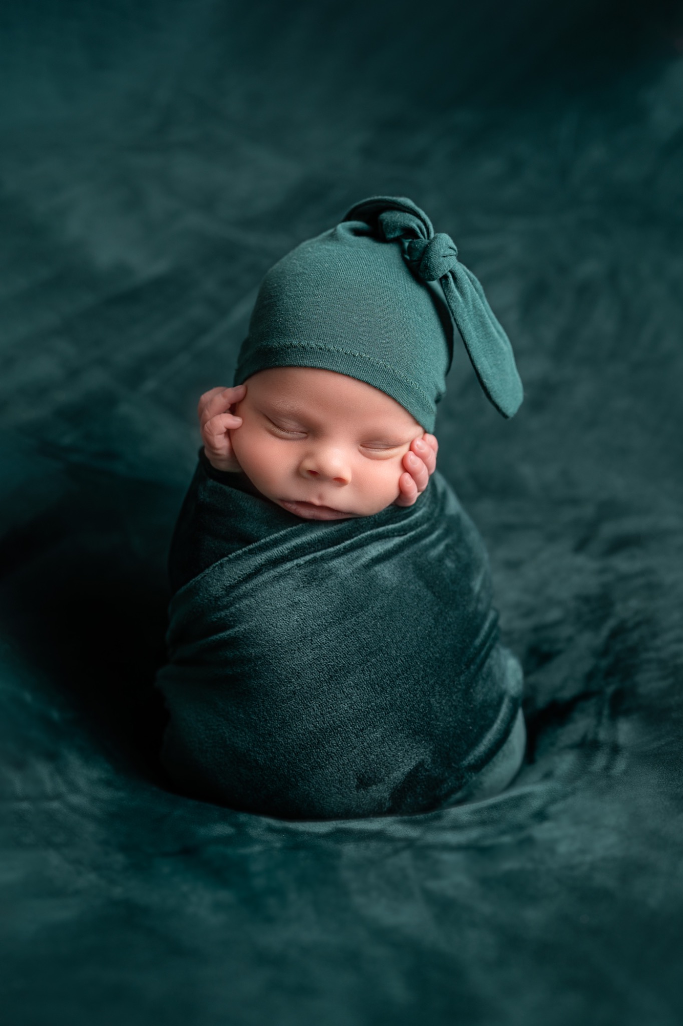 Śpiący noworodek owinięty w ciemnozielony, aksamitny materiał, z małą czapką z węzłem na głowie, leżący na dopasowanym, teksturowanym tle.